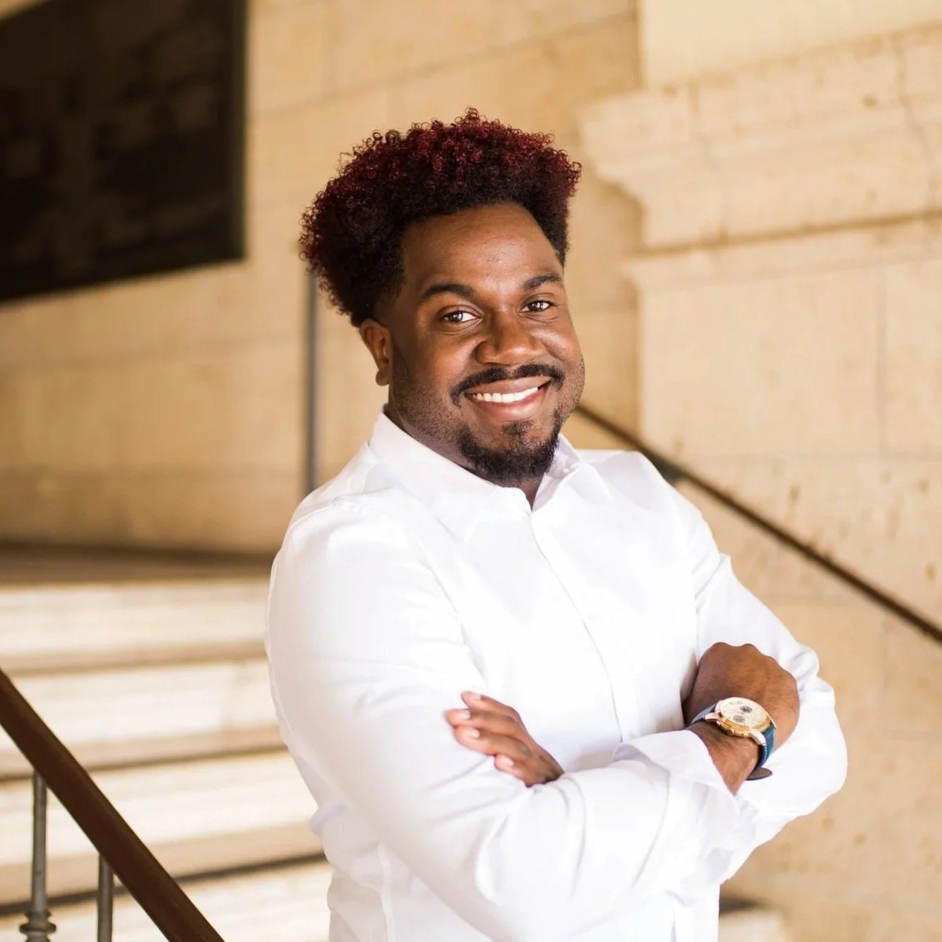 A smiling man with curly hair and a beard, wearing a white shirt and a watch, standing in front of a stone wall near a staircase.