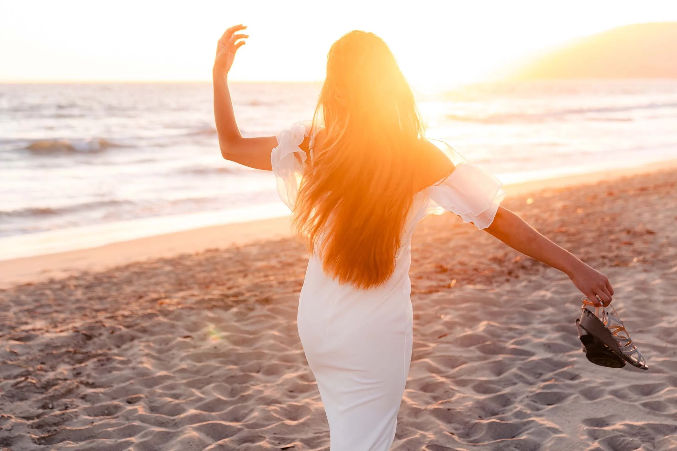 Woman holding shoes dancing on the beach at sunset