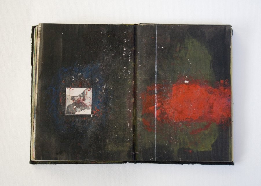 An open, weathered book with dark pages featuring abstract paint splatters in red, blue, and green. There is a small black-and-white photo of a person on the left page.