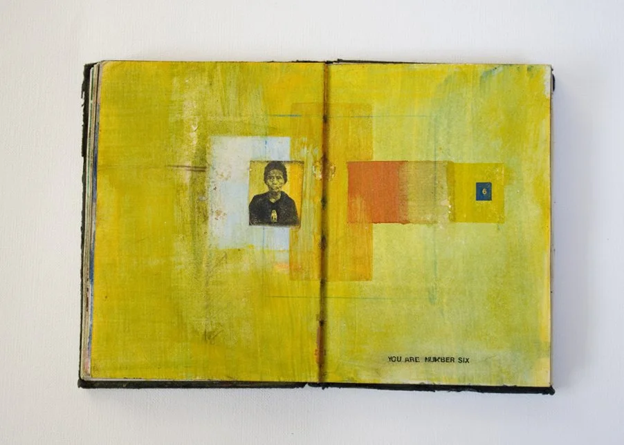 Open mixed media art journal with a yellow background, featuring a small black-and-white portrait of a man, a red rectangle, a yellow rectangle, and a blue square with the number 6, with black text at the bottom right saying "YOU ARE NUMBER SIX".