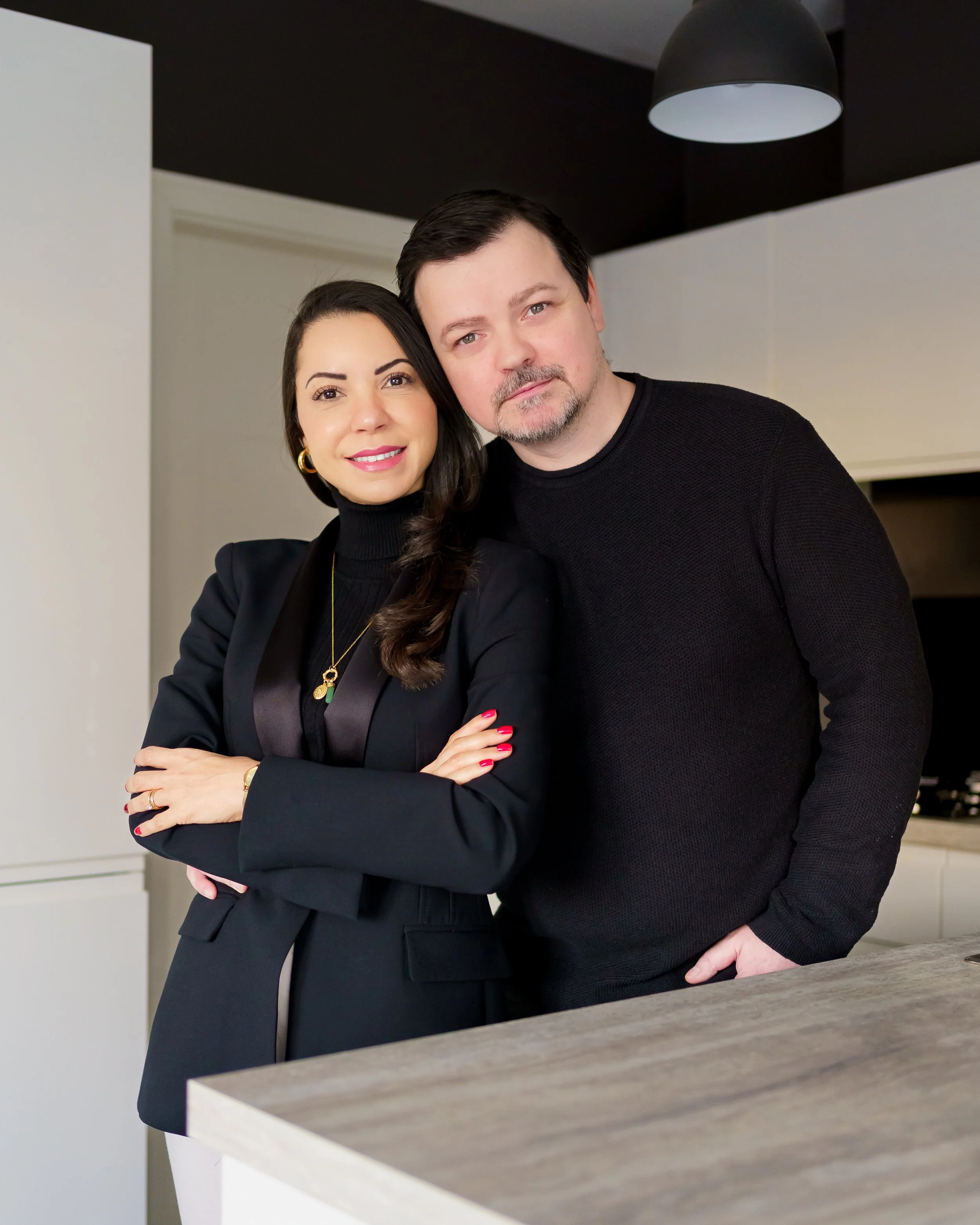 A woman with dark hair, wearing a black blazer and gold jewelry, standing next to a man with dark hair and a goatee, wearing a black sweater, in a modern kitchen.