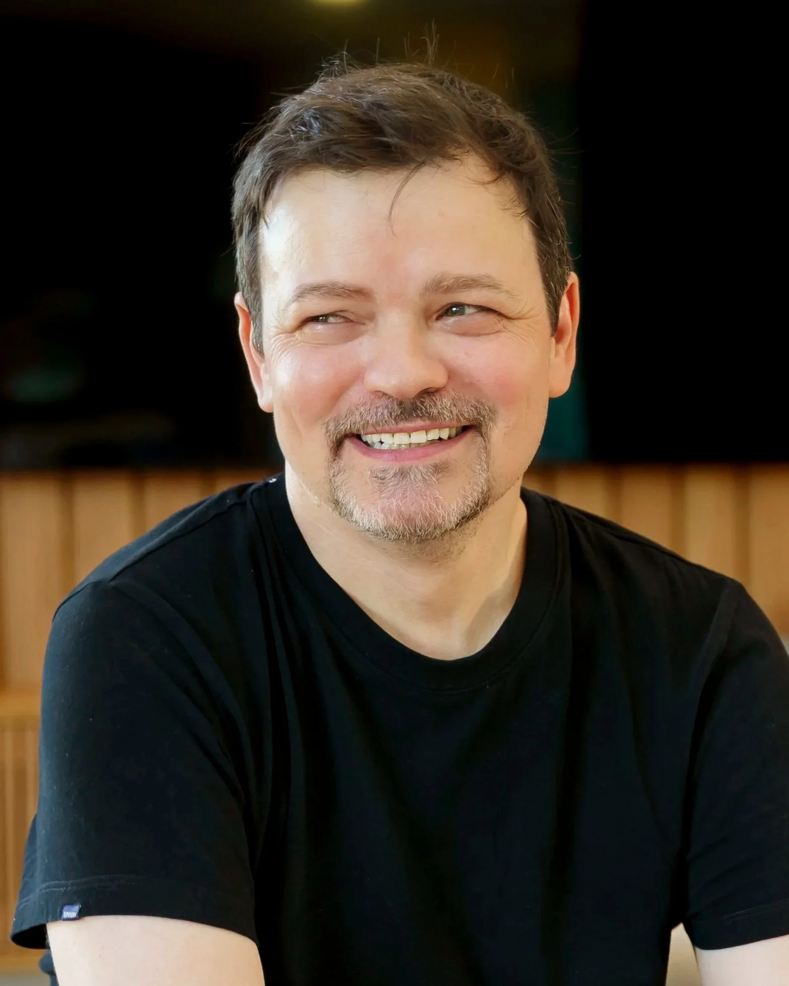 A man with light skin, brown hair, a mustache, and a goatee, smiling and looking to the side, wearing a black t-shirt, with a wooden wall in the background.