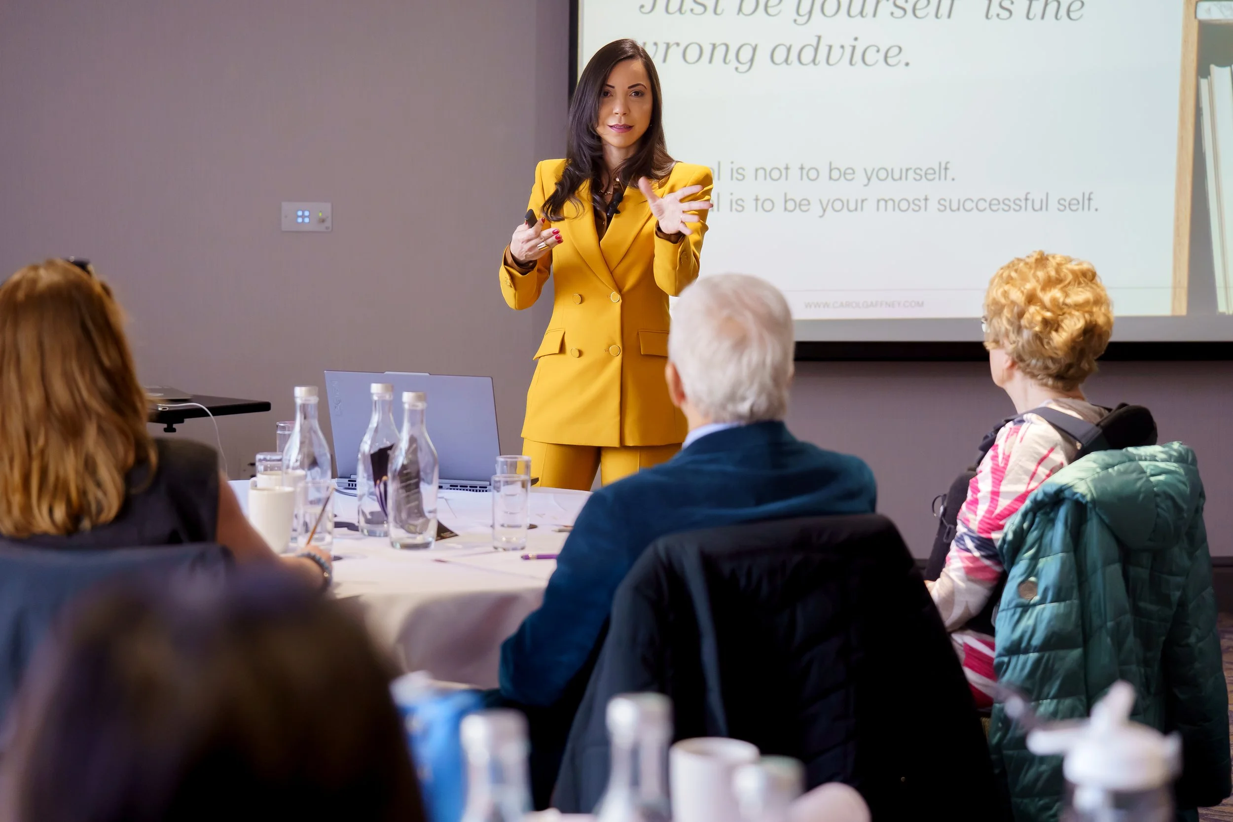 A woman in a yellow suit giving a presentation to a group of attendees seated at a table in a conference room, with a PowerPoint slide projected behind her.
