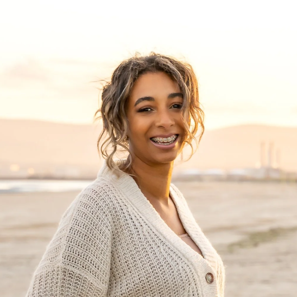 Young woman smiling at the beach during sunset, wearing a beige knitted cardigan and braces.