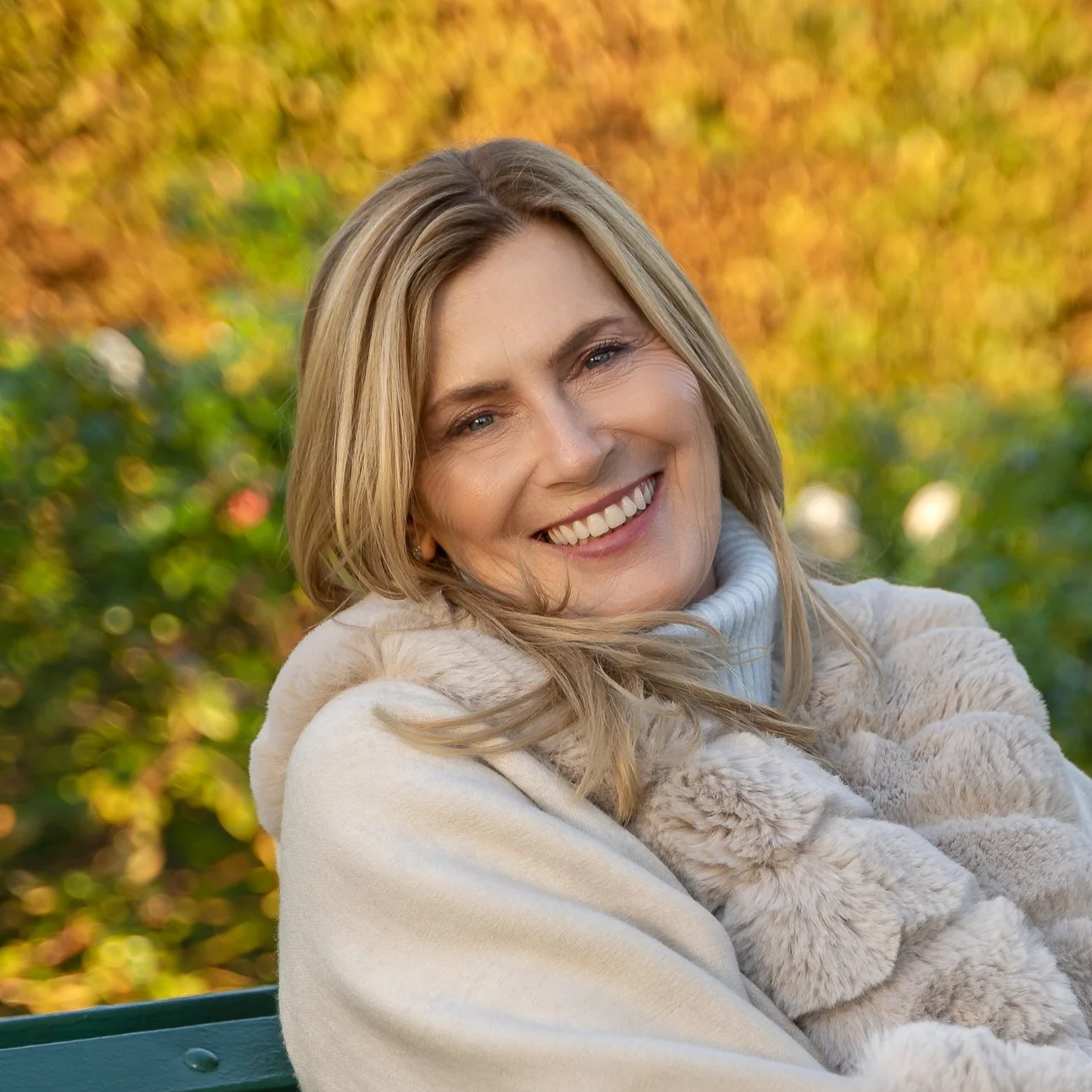 A woman smiling outdoors in autumn, wearing a beige fur coat and a white turtleneck, with fall foliage in the background.