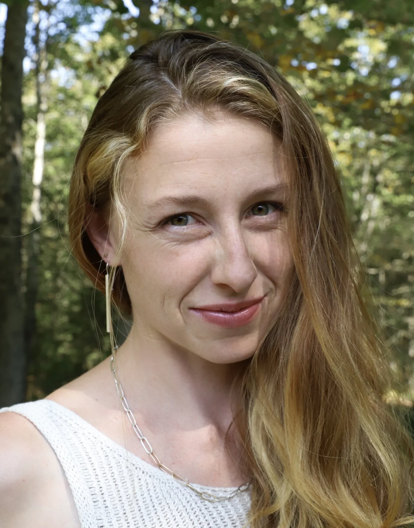 Woman with long, wavy red hair, green eyes, and a smile, wearing a knit top and a handmade silver paperclip chain necklace, and long light weight silver earrings in nature.