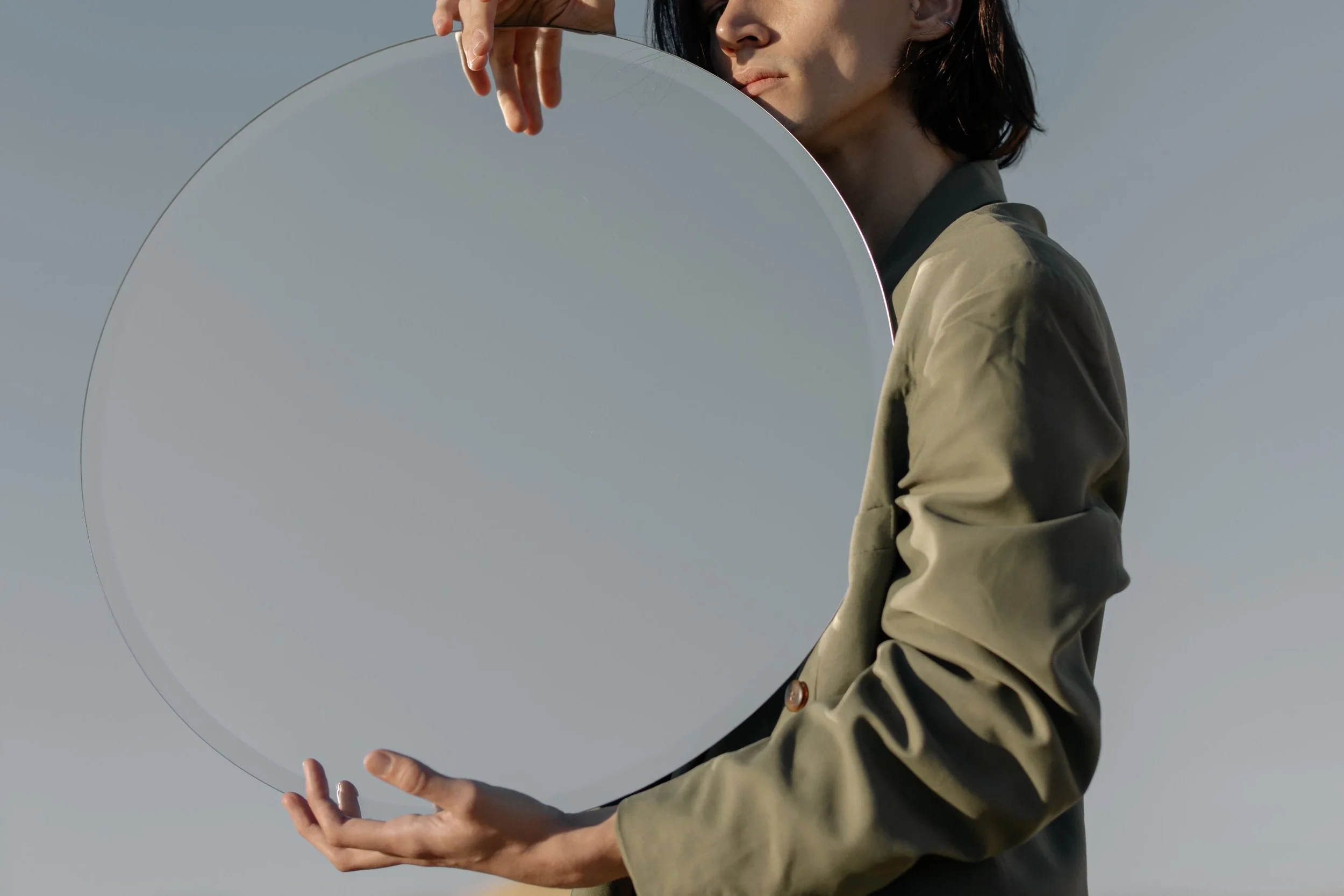A person with shoulder-length dark hair is holding a large round mirror in front of them, with part of their face visible as they look slightly to the side. The background is a plain, grayish sky.