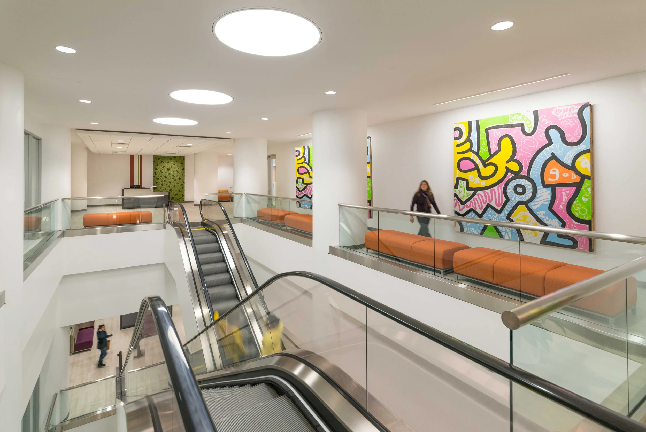 Modern indoor space with orange cushioned benches, abstract colorful wall art, escalator, and a woman walking.