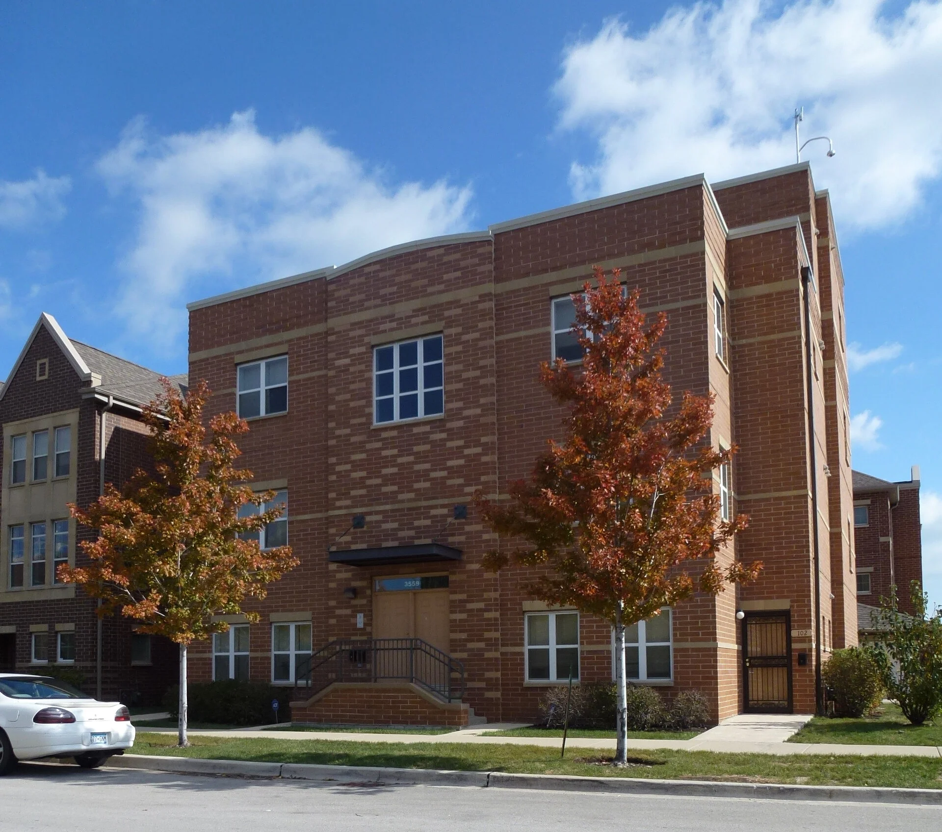 A multi-story brick apartment building with a small external staircase, surrounded by cars and trees with autumn foliage, under a blue sky with scattered clouds.