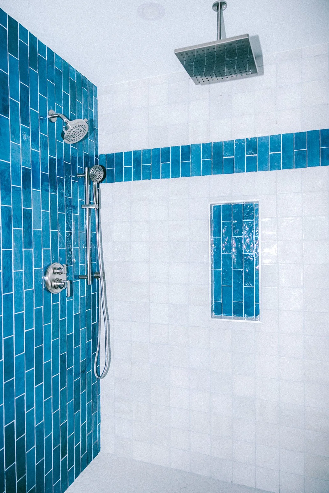 A modern shower with blue and white tiled walls, a large rain showerhead, a handheld shower with a sliding bar, a built-in niche with blue tiles, and a ceiling-mounted square rain shower.
