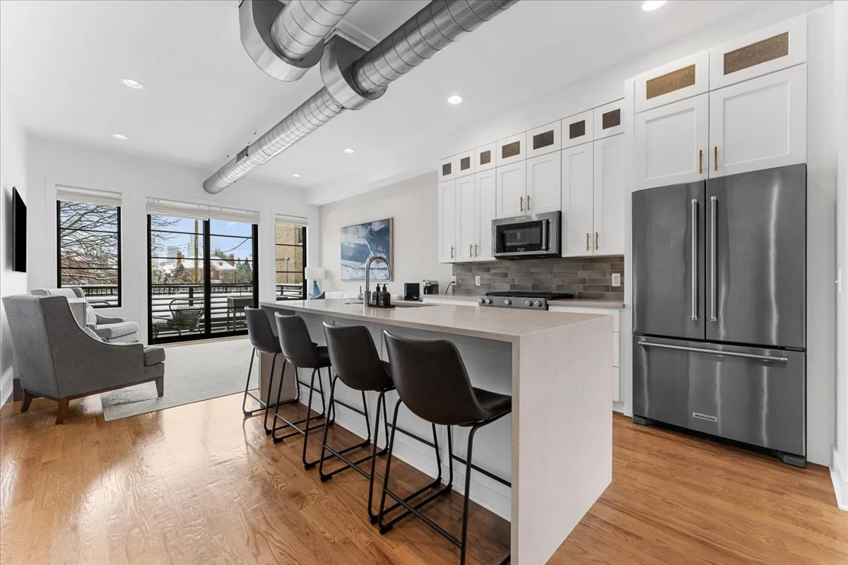 Modern kitchen with white cabinets, stainless steel refrigerator, black microwave, gray backsplash, and a kitchen island with four black chairs, adjacent to a living area with chairs and large windows.