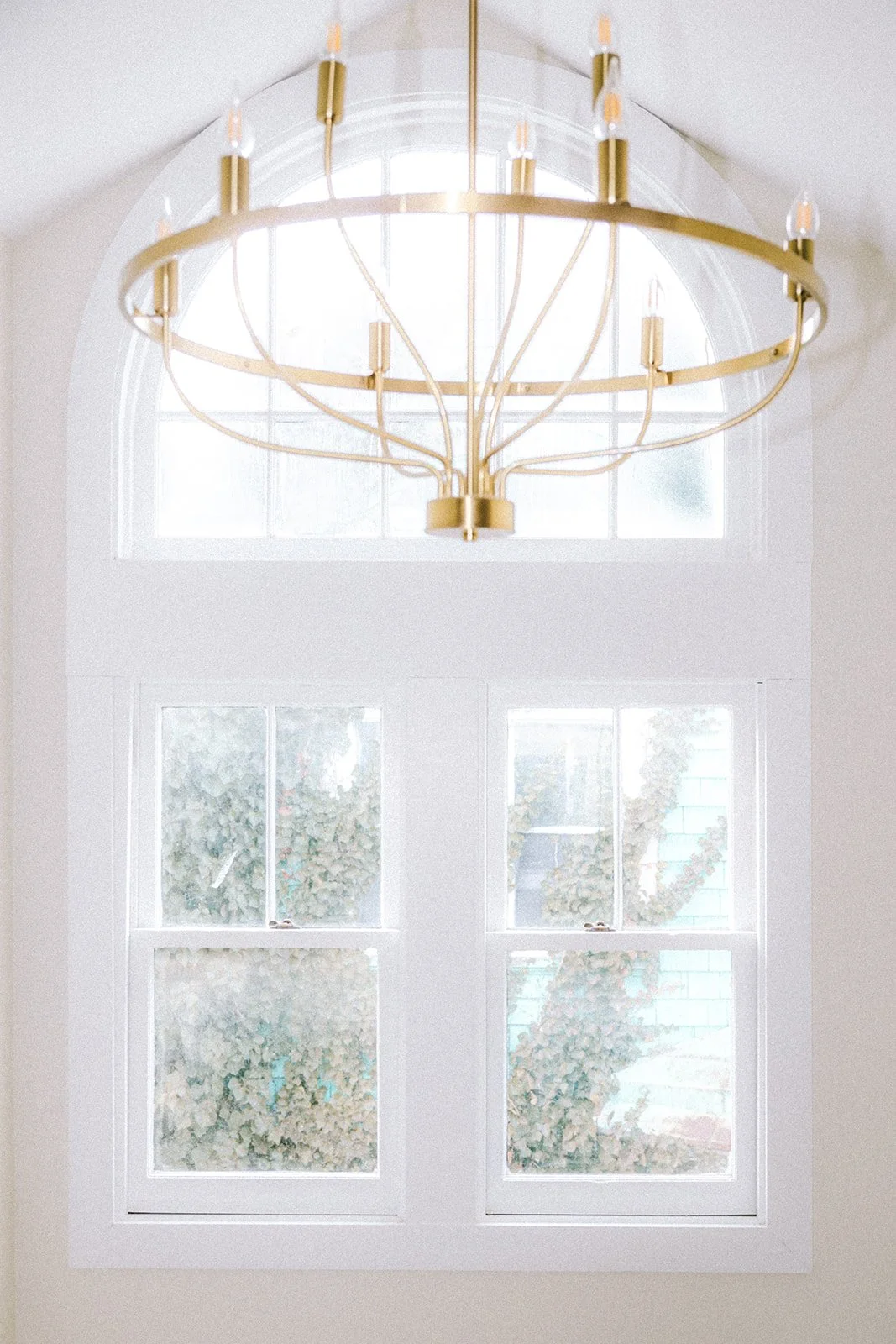 An elegant gold chandelier hanging above two white-framed windows with frosted glass and greenery outside.