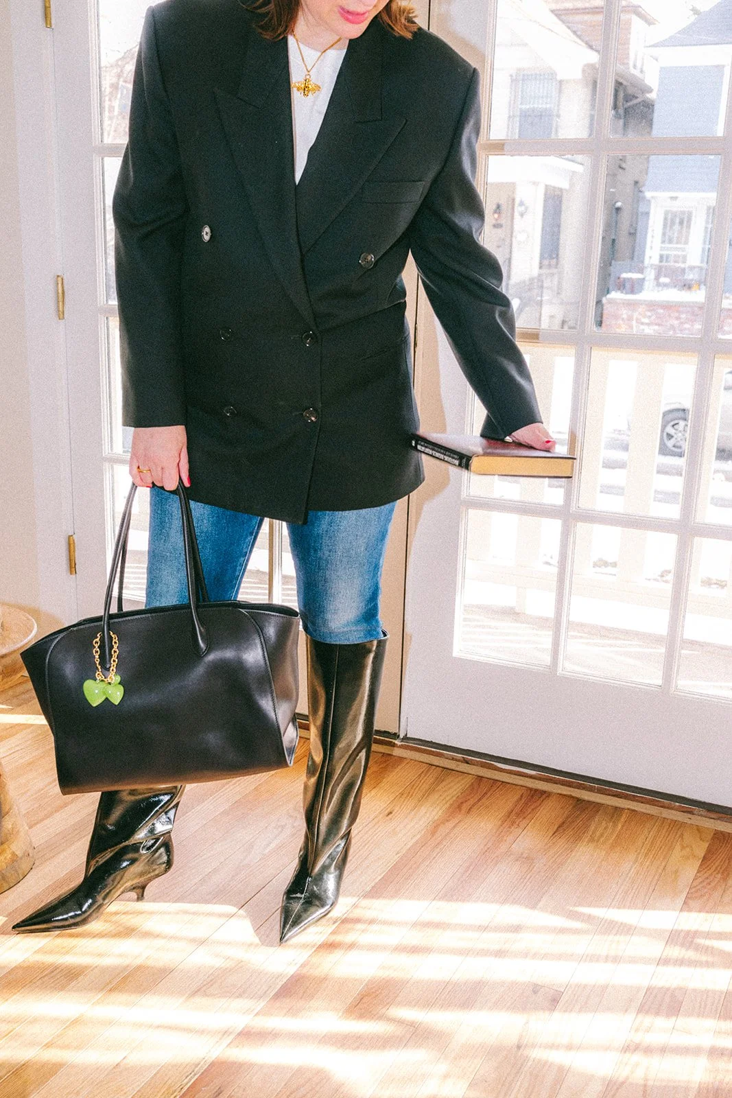 Person wearing black blazer, denim jeans, and shiny black boots, holding a large black handbag with a green butterfly keychain, standing near a glass door with sunlight streaming in.