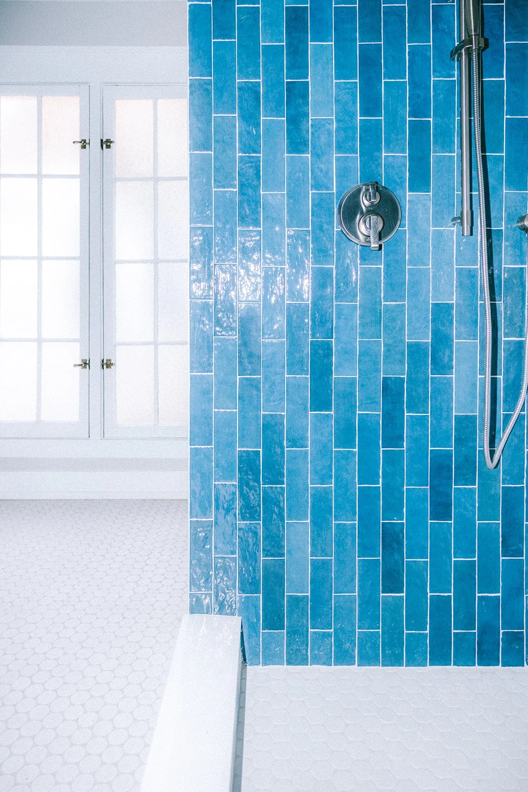 Close-up of a bathroom shower area with blue rectangular tiles, a round silver shower control, and a handheld showerhead on a vertical bar, with a window in the background.