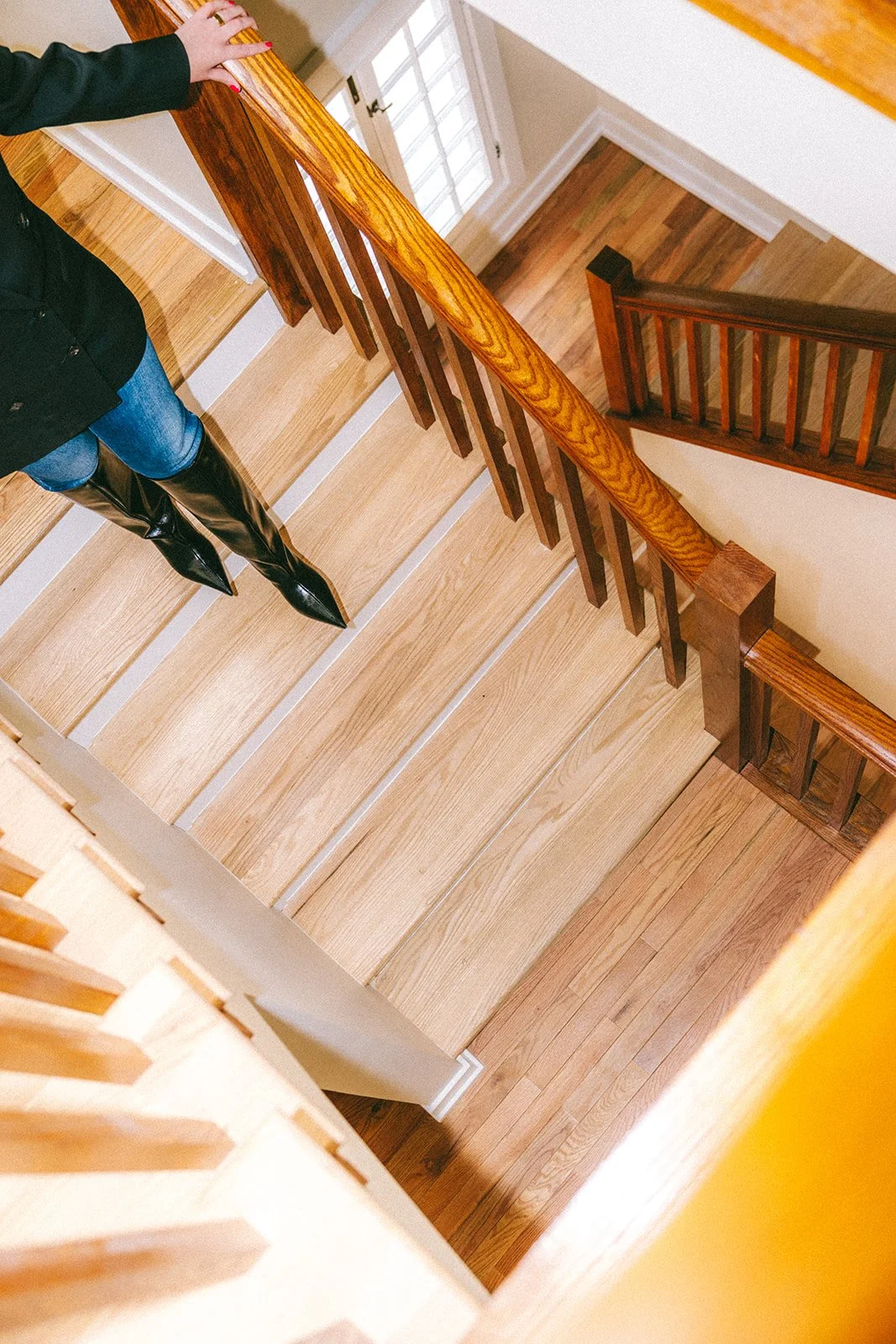 Top-down view of a wooden staircase with a person standing on it, holding the railing. The person is wearing black boots, blue jeans, and a black jacket. The stairway has light wood steps and dark wooden railings, with a small landing at the top near