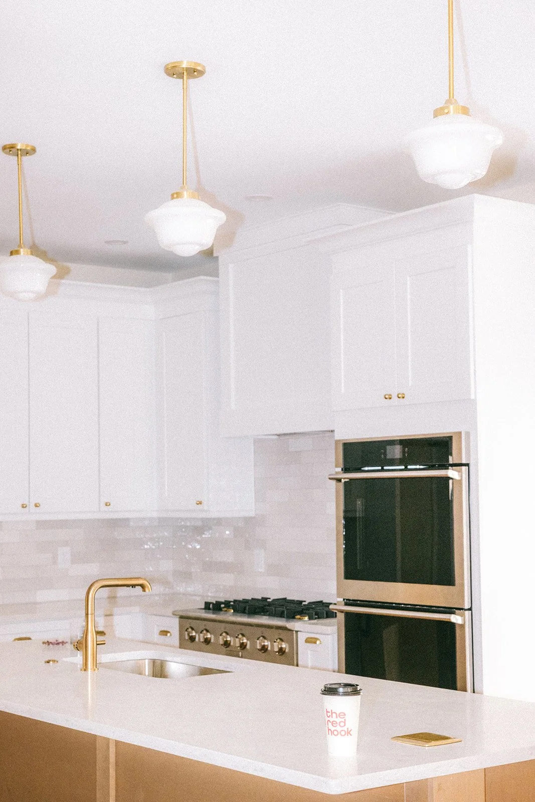 Bright white kitchen with gold accents, ceiling lights, double oven, gas stove, gold faucet, coffee cup, and countertop.