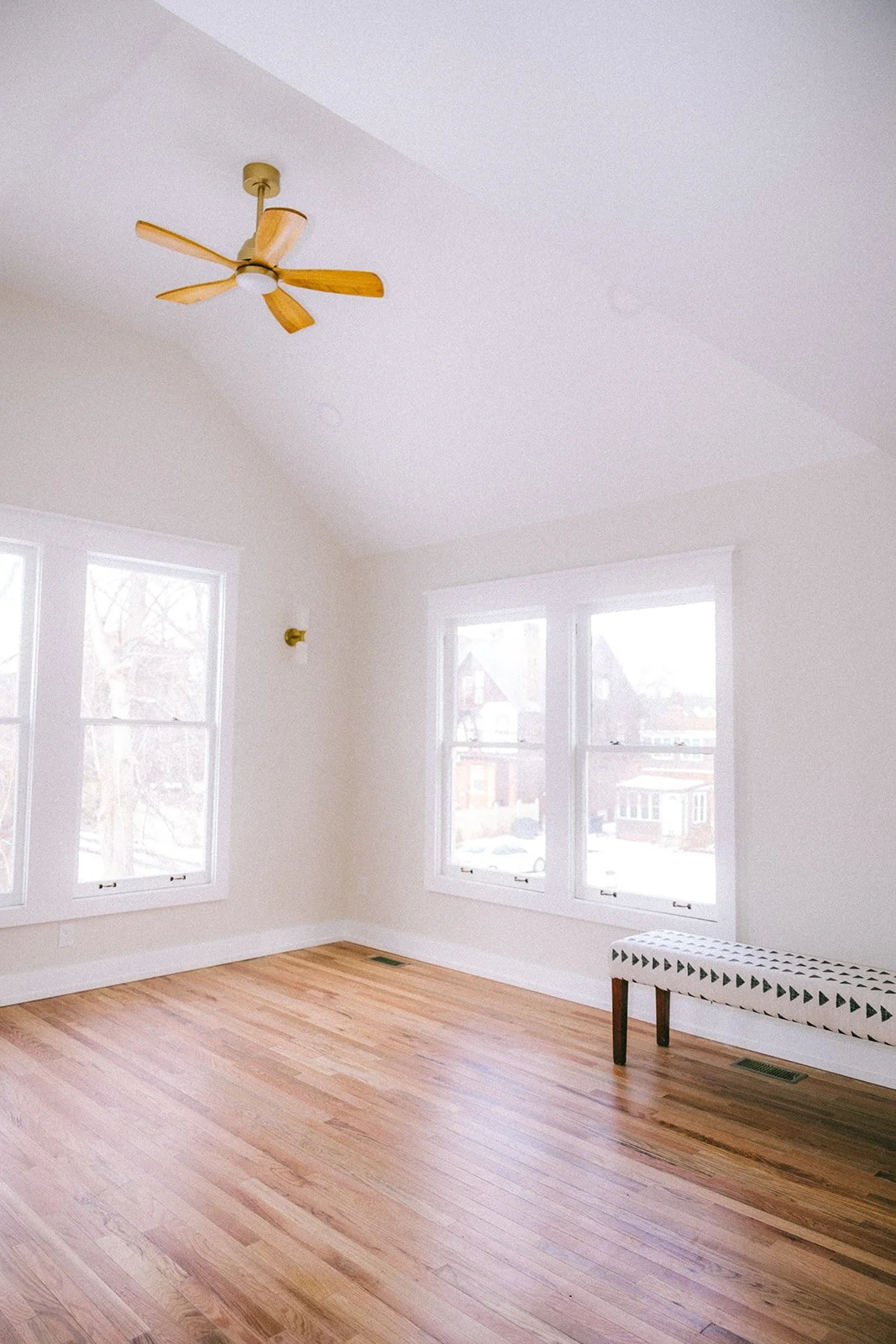 Empty room with white walls, wooden floor, large windows, ceiling fan, and a black and white patterned bench.