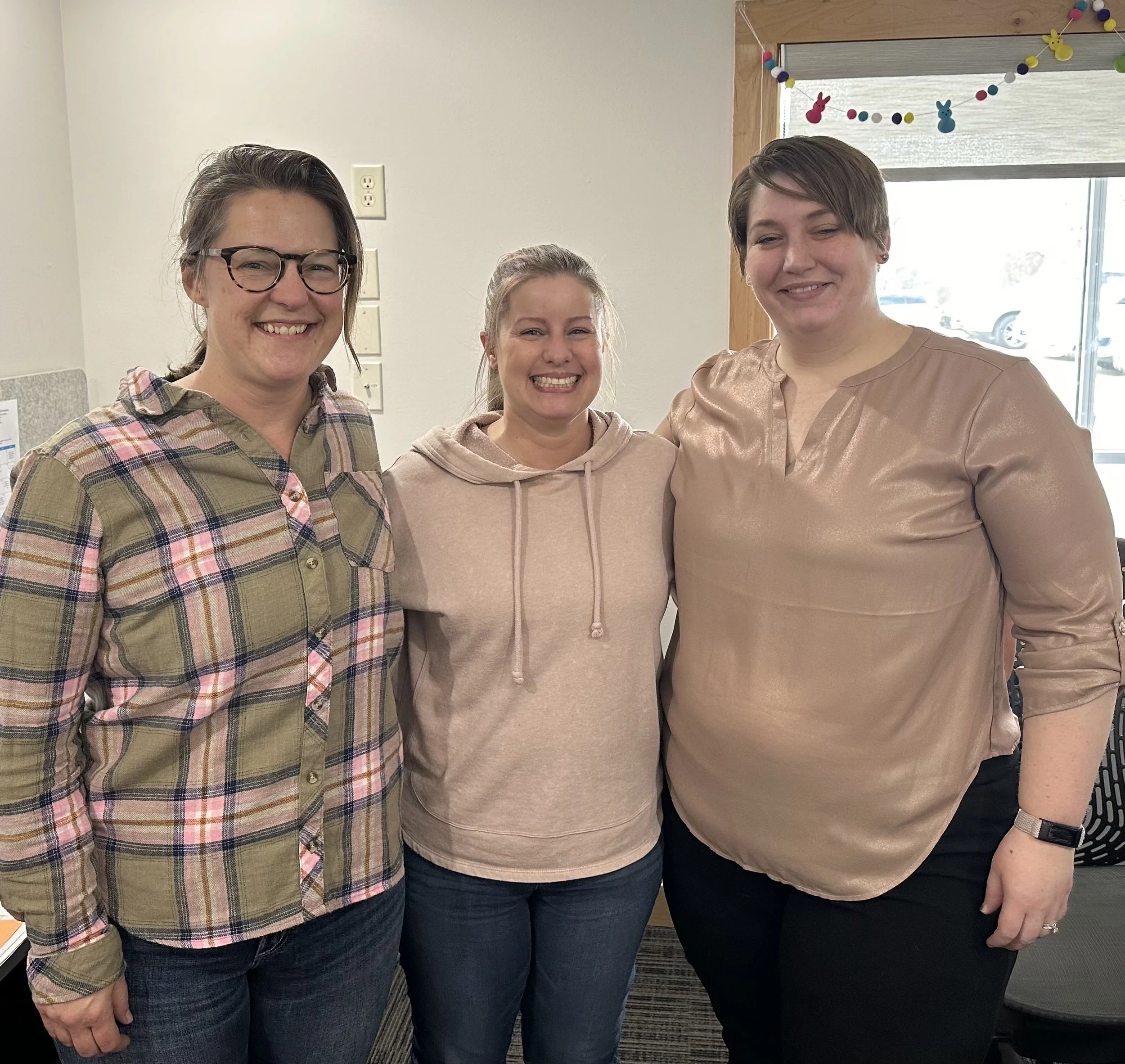 Three women smiling and standing close together indoors near a window decorated with colorful paper bunny and egg garland.