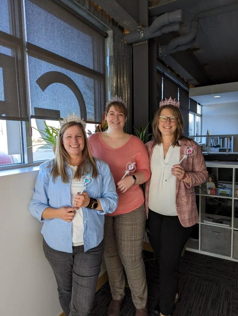 Three women wearing tiaras standing indoors, holding wands, smiling for a celebration event.