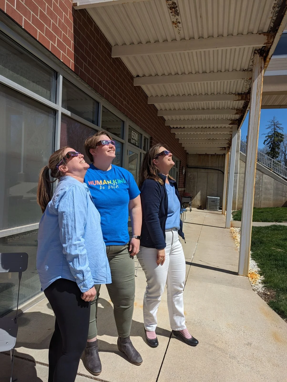 Three women standing outdoors under a covered walkway, looking up and smiling, with sunlight on their faces.