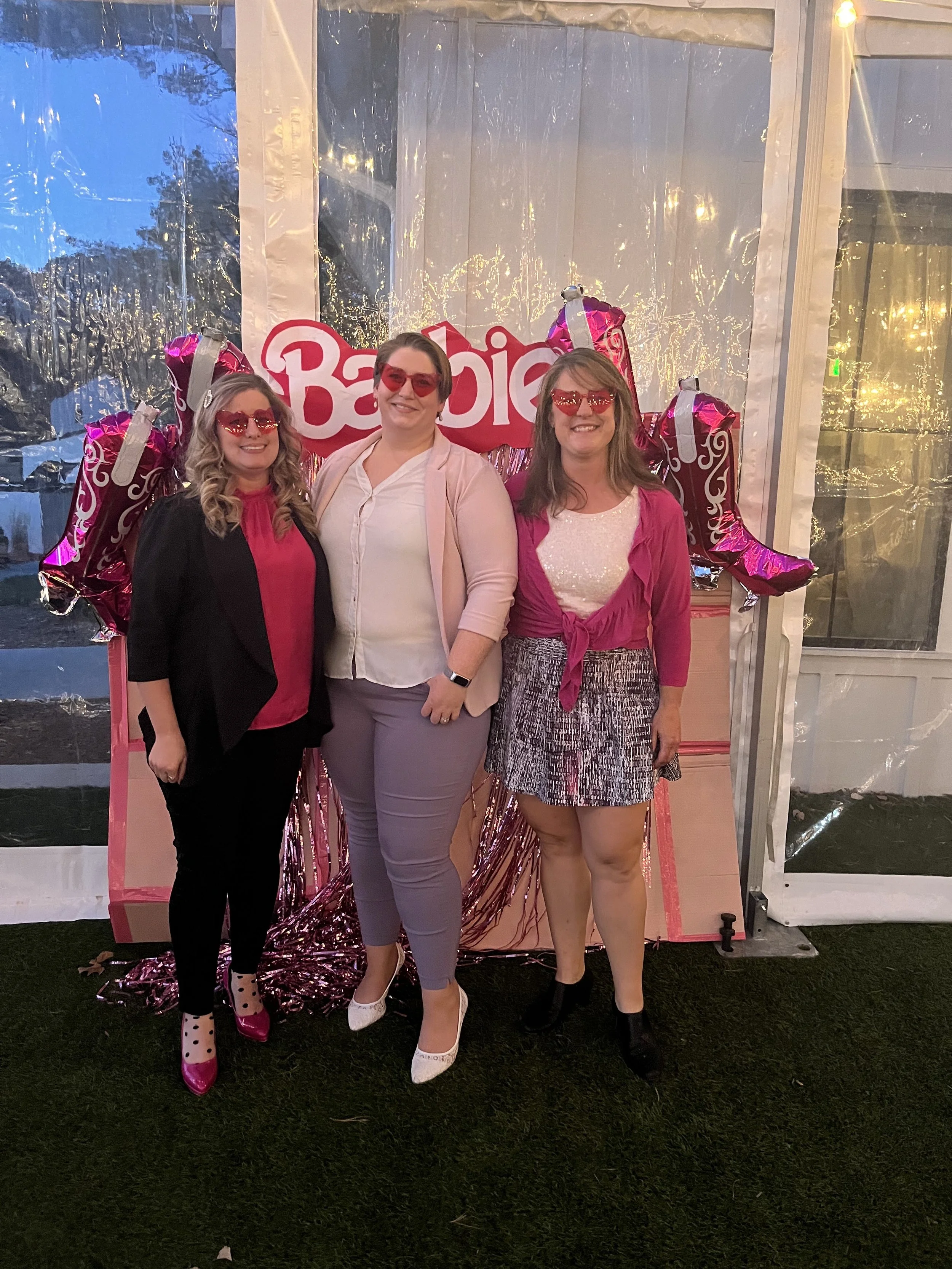 Three women standing in front of a Barbie-themed backdrop with pink balloons that spell 'Barbie.' They are all wearing pink and black clothing and pink sunglasses, smiling at the camera.