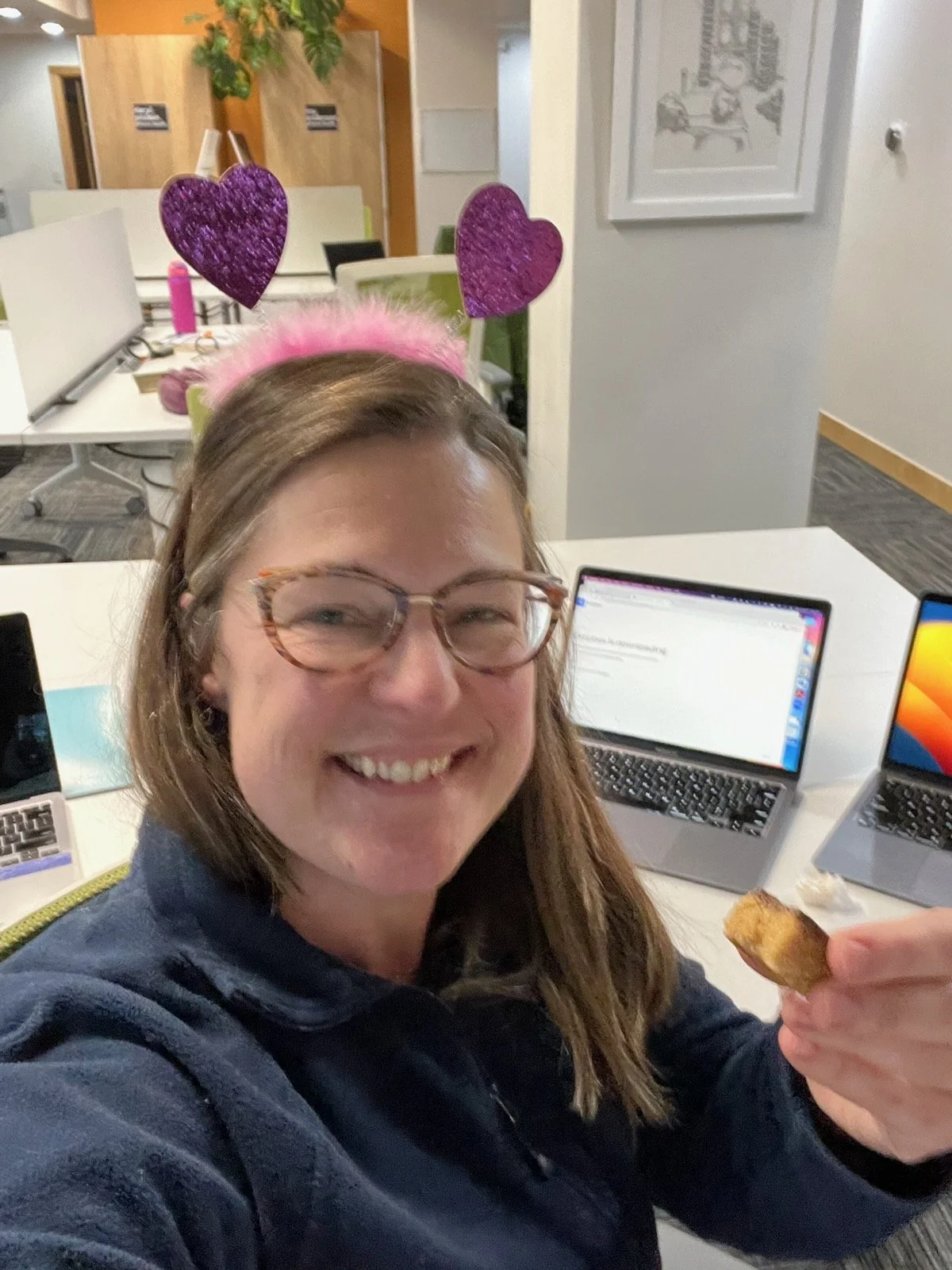 A woman wearing pink heart-shaped headband with heart antennas, smiling, holding a snack, sitting at a table with open laptops in an office setting.