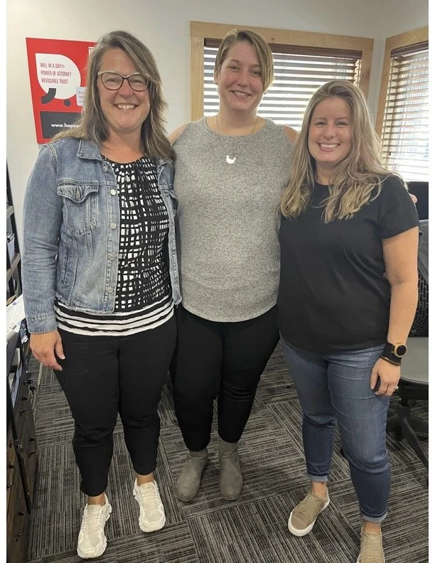 Three women standing together indoors, smiling at the camera. The woman on the left wears glasses, a denim jacket, and black pants. The woman in the middle wears a gray top and black pants. The woman on the right wears a black t-shirt and jeans, with