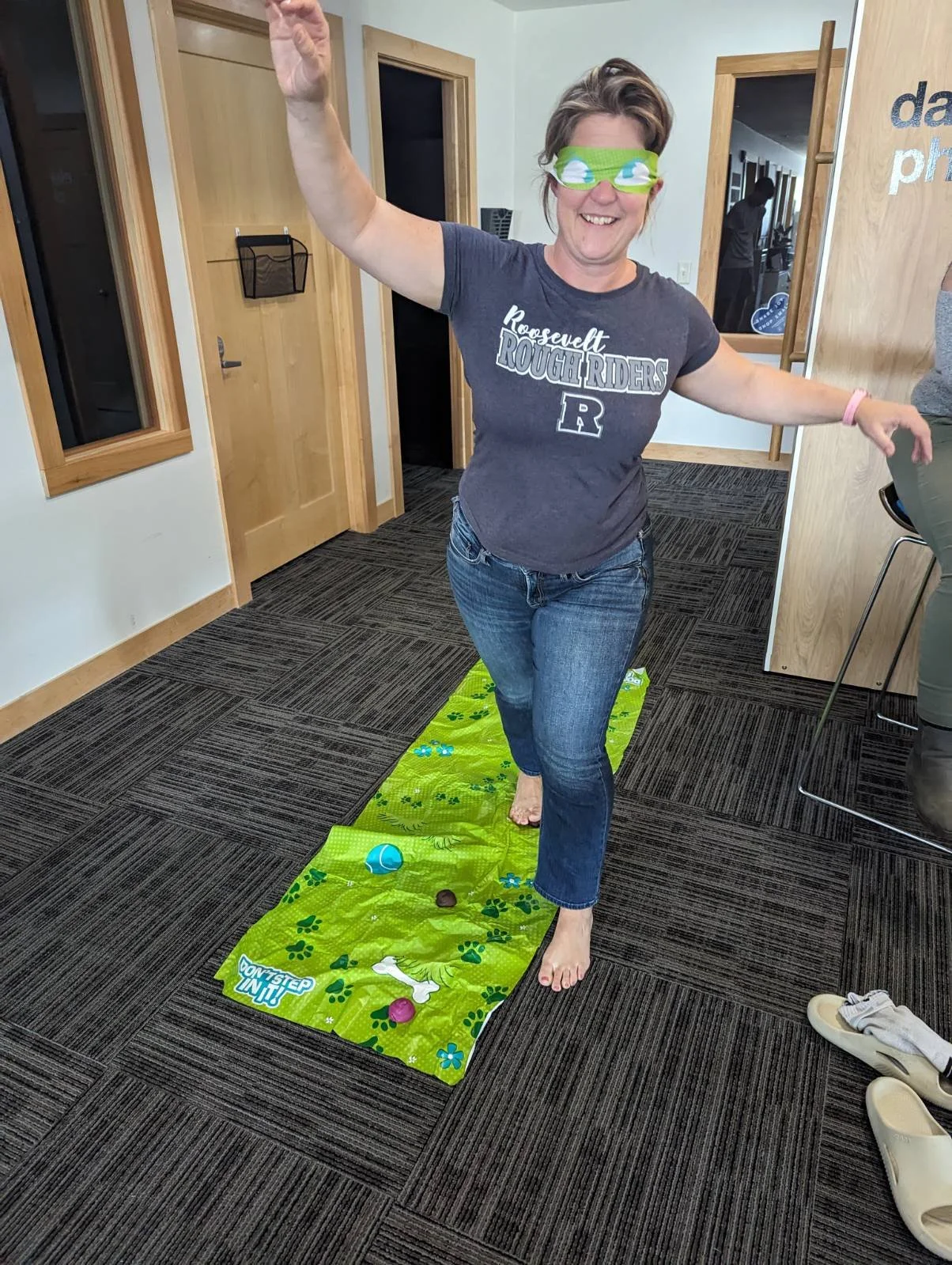 A woman wearing a Roosevelt Rough Riders t-shirt and a green blindfold is playing a game of indoor bean bag toss while walking on a green mat with paw print designs.