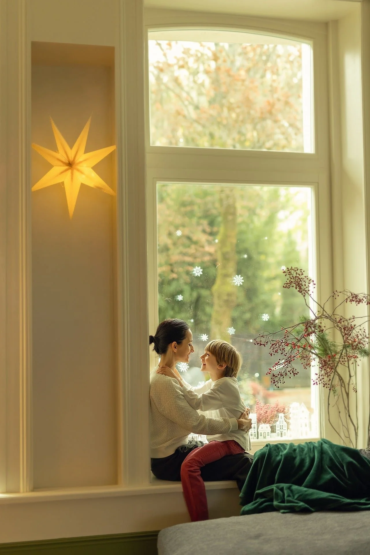 A woman and a young boy are sitting together on a window sill, facing each other and smiling. The scene is decorated for winter with snowflake window stickers and a lit star-shaped ornament hanging inside the window.