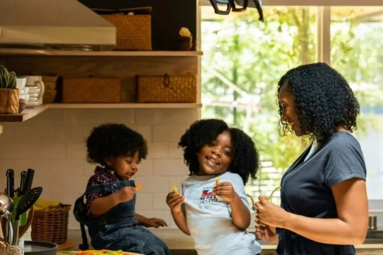 A woman and two children with curly hair smiling in a kitchen, preparing snacks and engaging with each other.