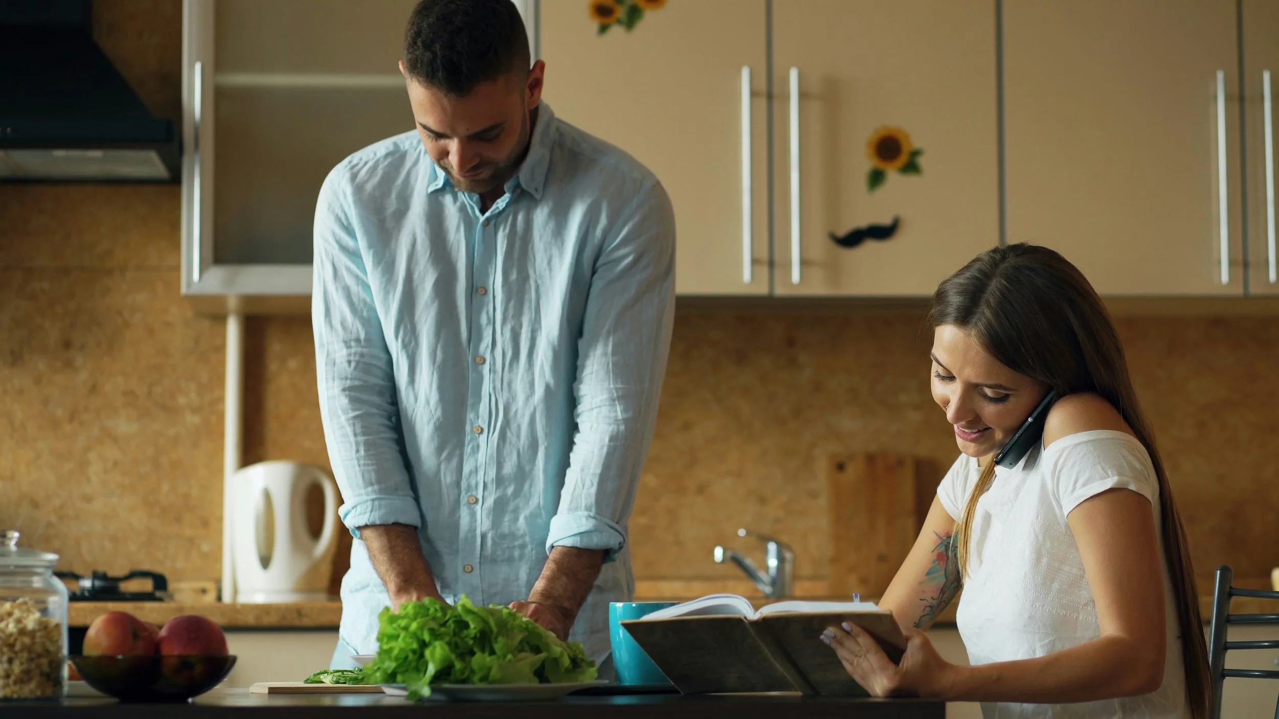 A man and a woman in a kitchen, with the woman talking on the phone while reading a recipe or note, and the man chopping vegetables on the counter.