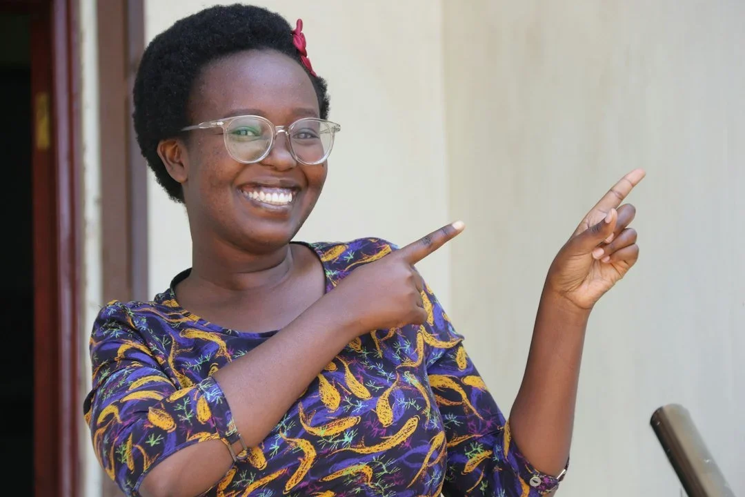 A smiling woman with glasses pointing to the right, wearing a patterned dress, in front of a plain wall.