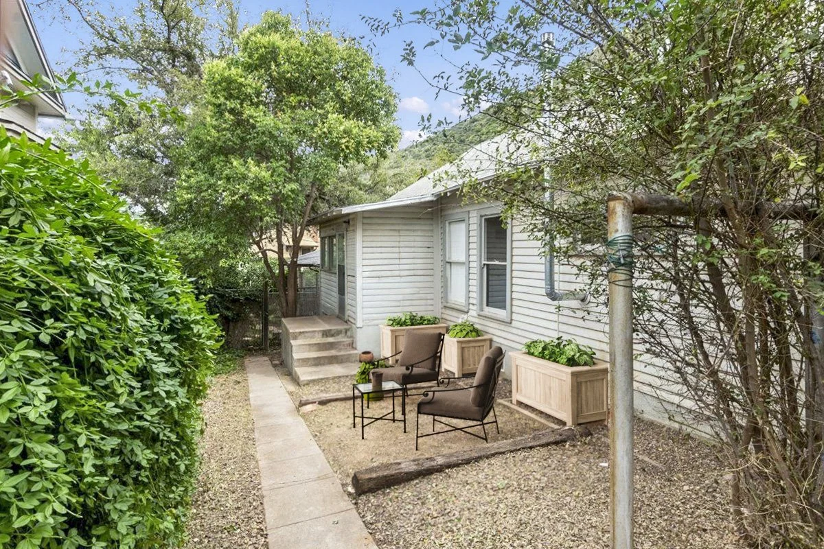 Small backyard with a concrete pathway, two chairs with side table, potted plants, and surrounding bushes and trees.