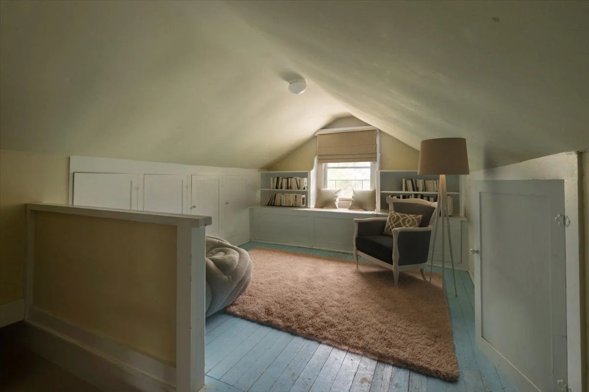 Attic bedroom with built-in white storage, a window seat with pillows, a beige rug, a black armchair with a patterned pillow, a floor lamp, and bookshelves on either side of the window.