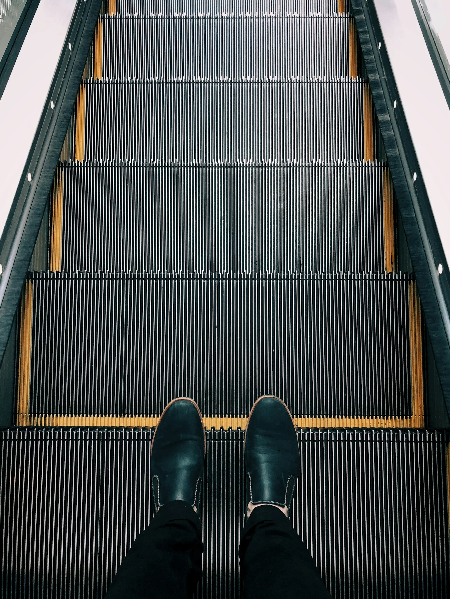 View from someone's perspective standing on an escalator, wearing black shoes and black pants, with the escalator moving upward.