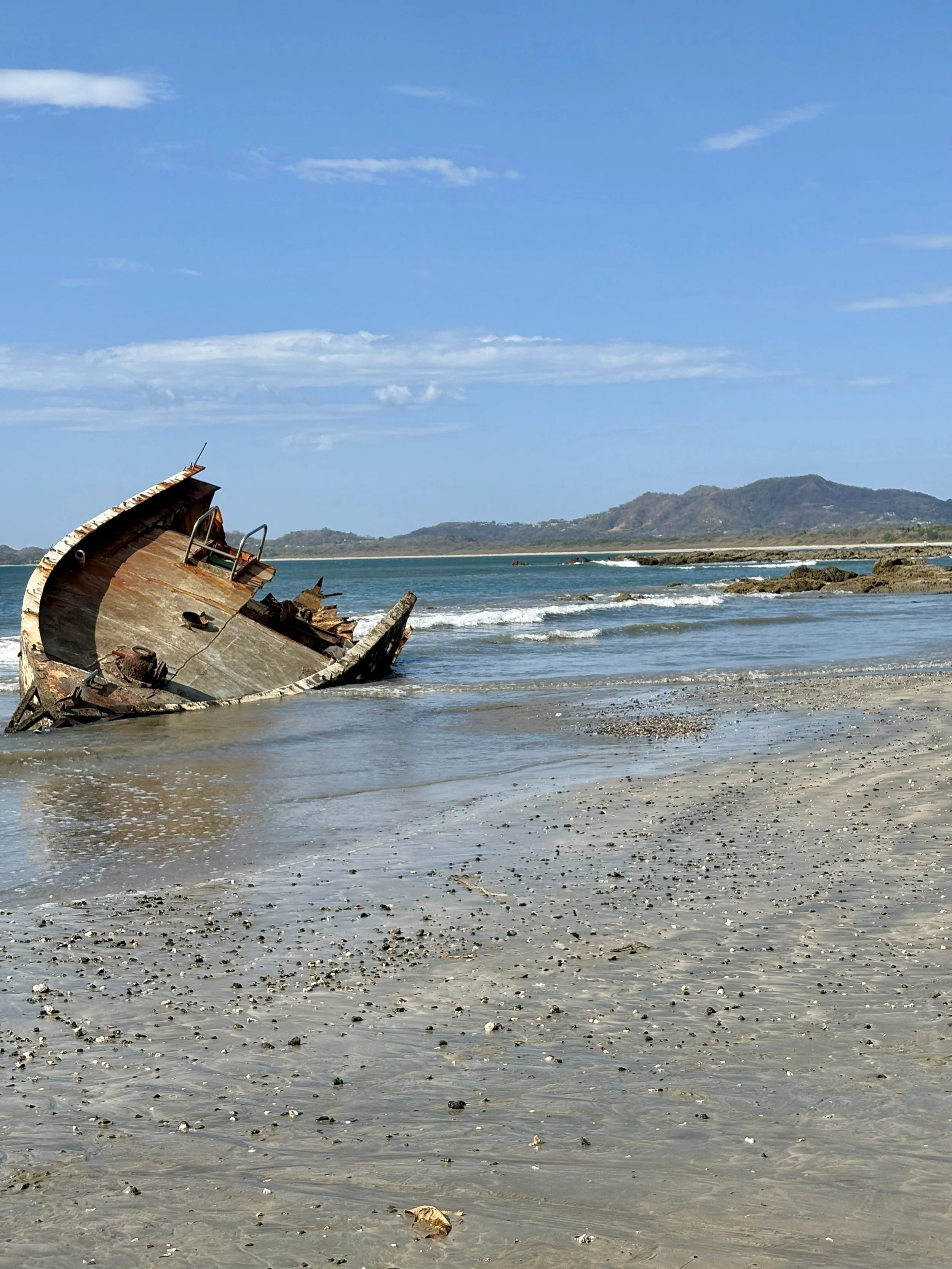 A rusted, sunken shipwreck partially submerged on a sandy beach with small waves and mountains in the background.