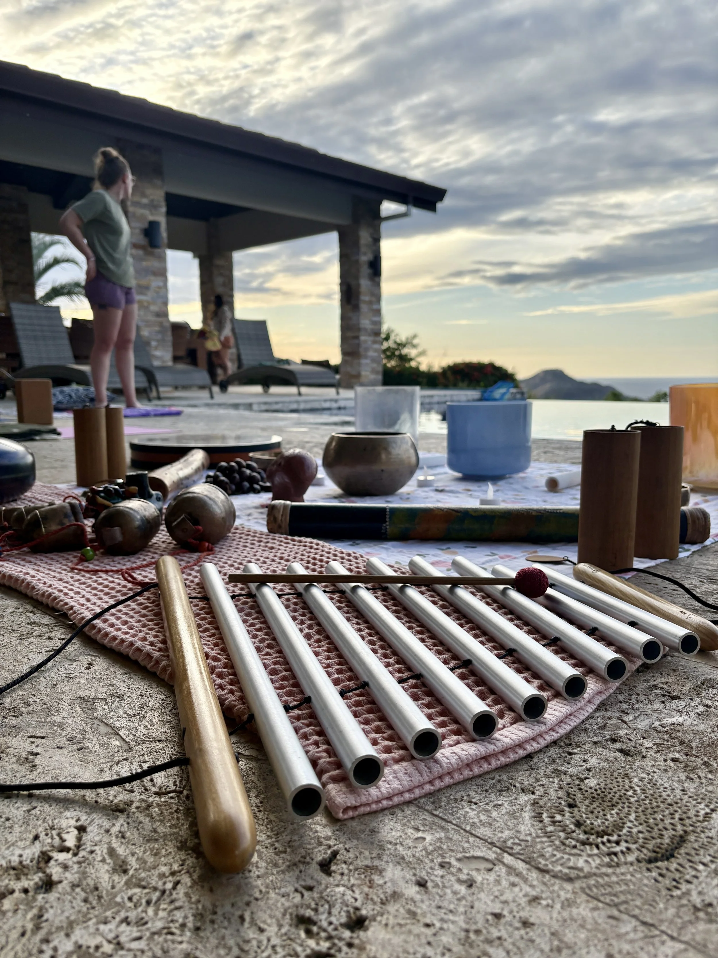 Latin percussion instruments on a pink mat, including a wooden agogo, metal tubes, and a mallet, set outdoors near a pool at sunset with people practicing yoga in the background.