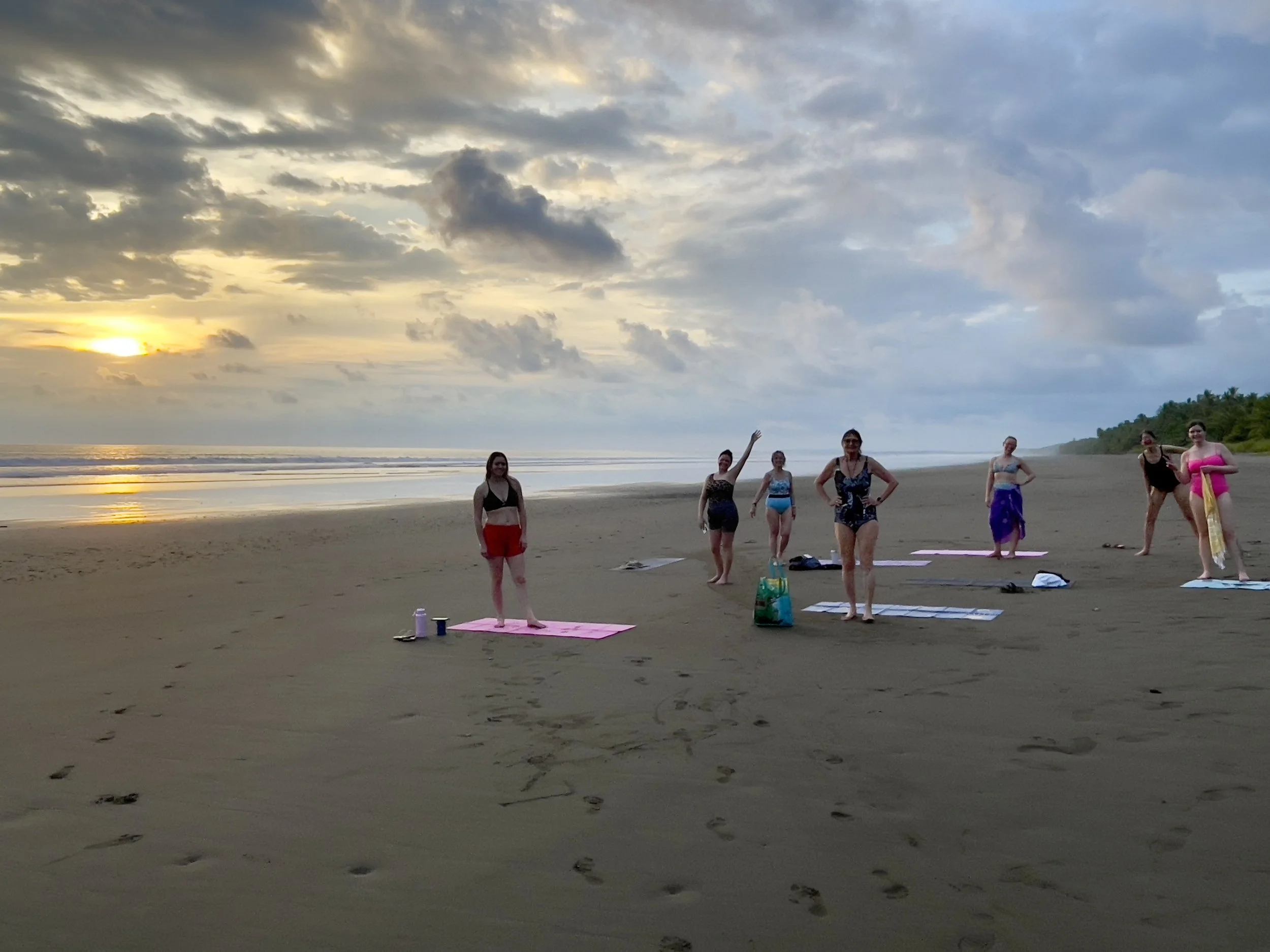 A group of women practicing yoga on a beach during sunset, with cloudy sky and ocean waves in the background.