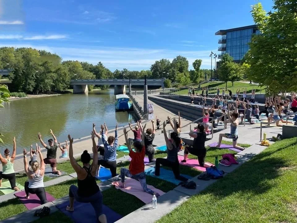 Group of people practicing yoga outdoors on a sunny day near a river with trees and a modern building nearby.