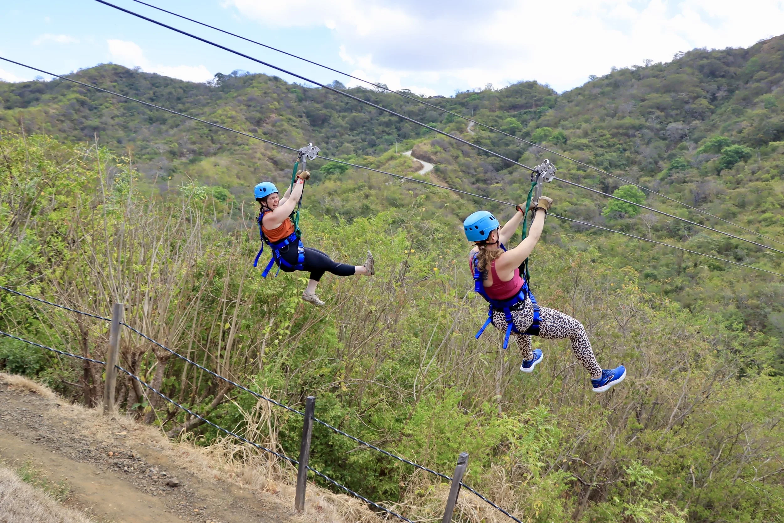 Two women zip lining through a green mountainous landscape, wearing helmets and safety harnesses.