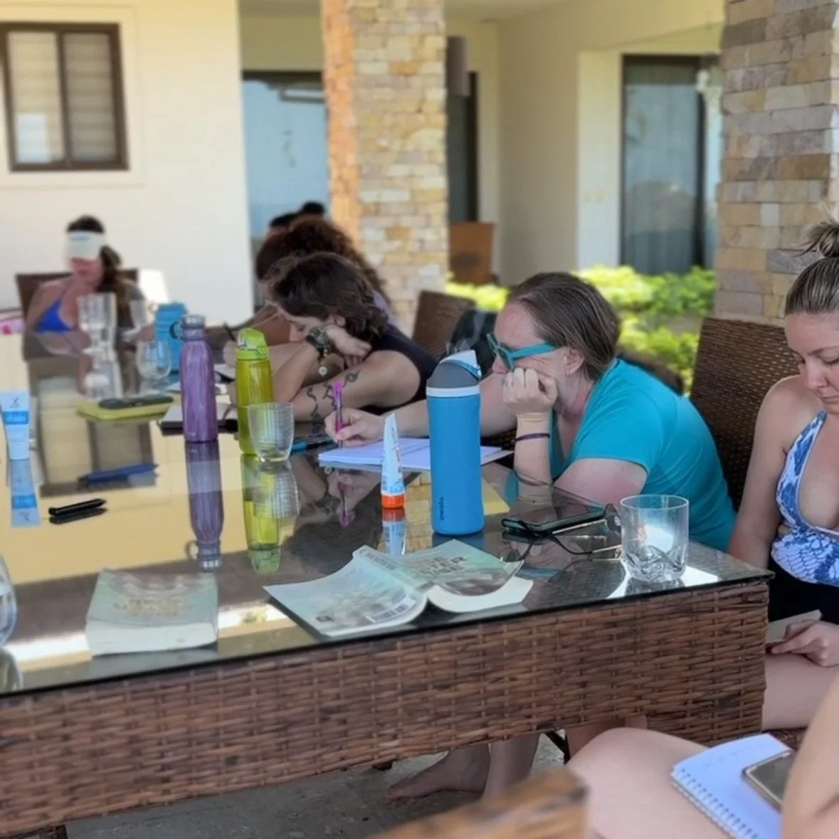 Group of women sitting at a table, appearing bored or tired, with notebooks, water bottles, and glasses on the table in an outdoor patio setting.