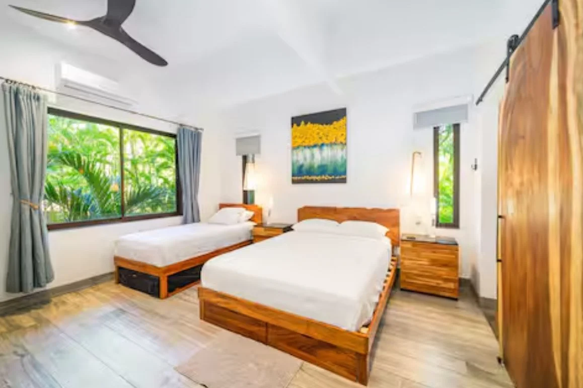 A bedroom with two beds, large window with greenery outside, wooden furniture, and modern décor