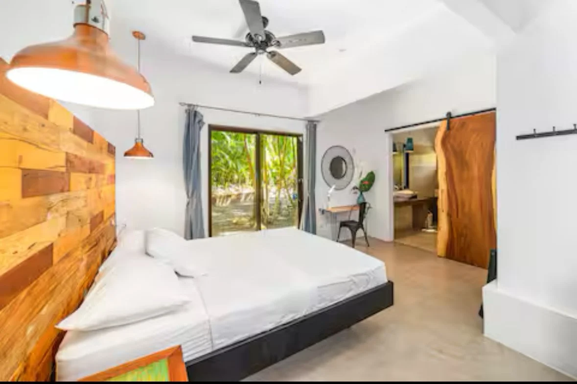 Modern bedroom with a large bed, wooden headboard, ceiling fan, sliding glass door with curtains, desk area with a mirror, and for sliding barn doors, one open revealing an interior space.