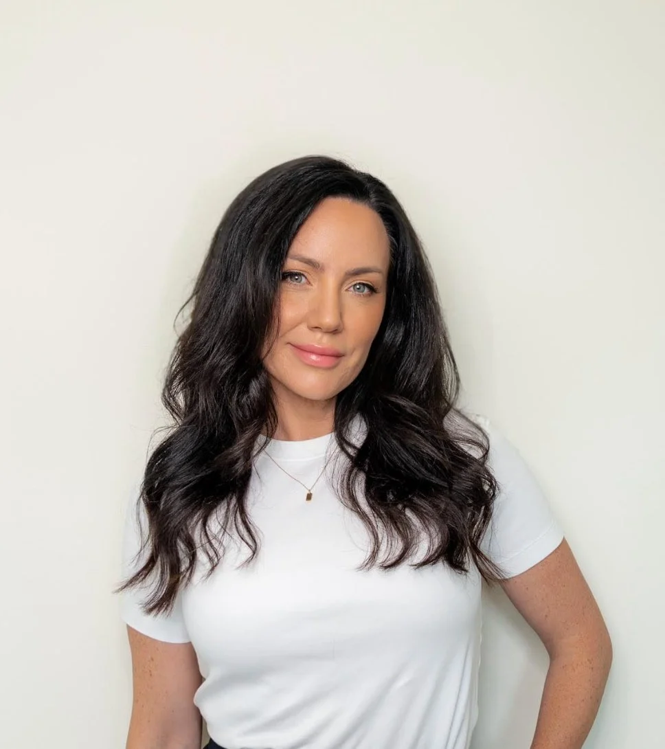 Woman smiling with dark brown wavy hair. She is wearing a white t-shirt and a small gold chain.