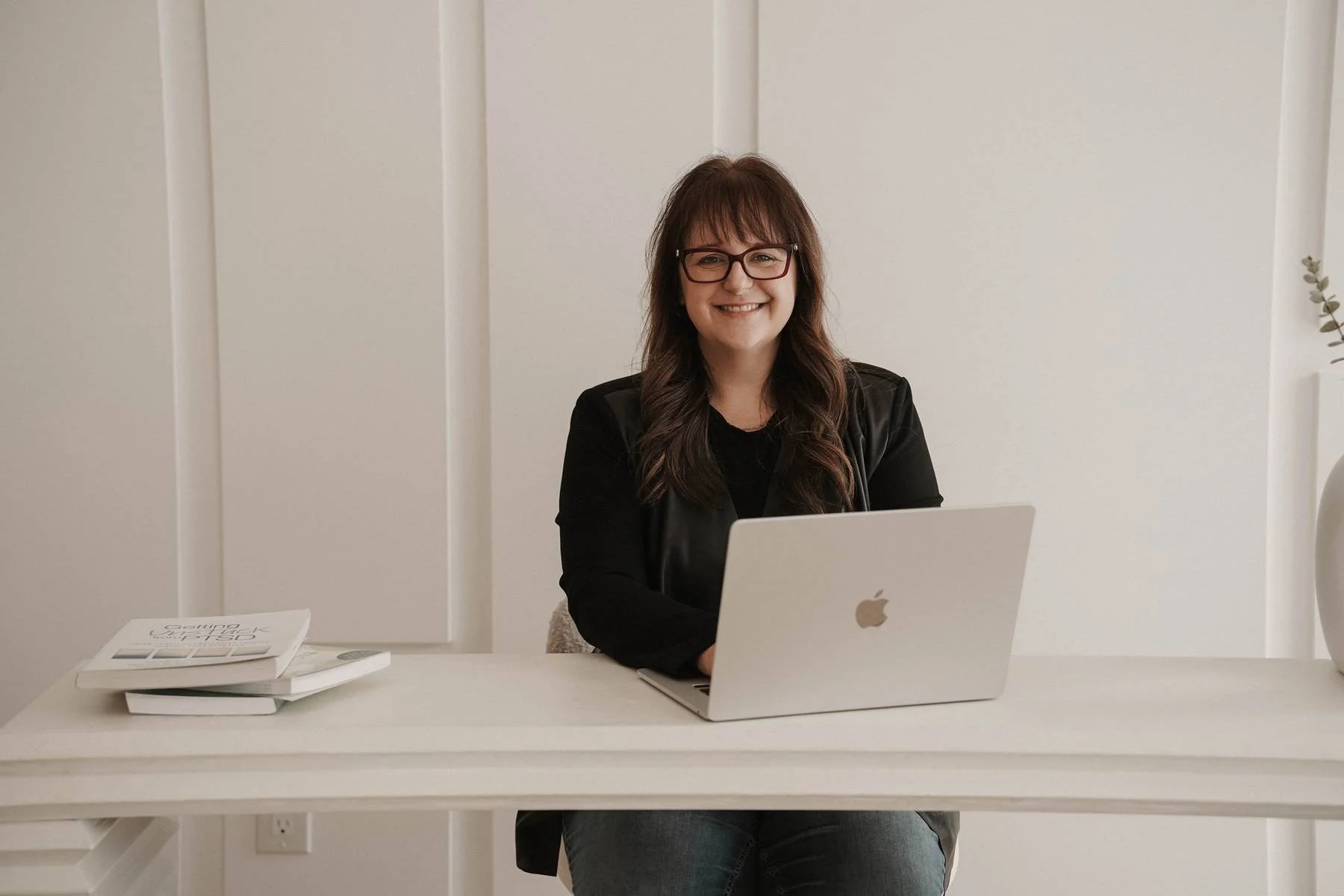 A woman with glasses sitting at a white desk with a silver laptop and some books, smiling at the camera in a minimalist room.