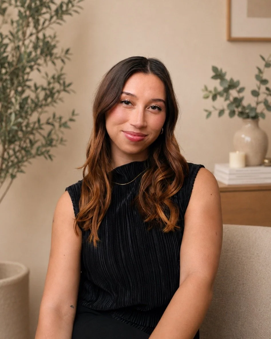 A young woman with long, wavy brown hair, wearing a black sleeveless top, sitting indoors with a neutral expression. In the background, there is a large potted plant, a framed picture, and a wooden table with a stack of books, a candle, and a white vase.
