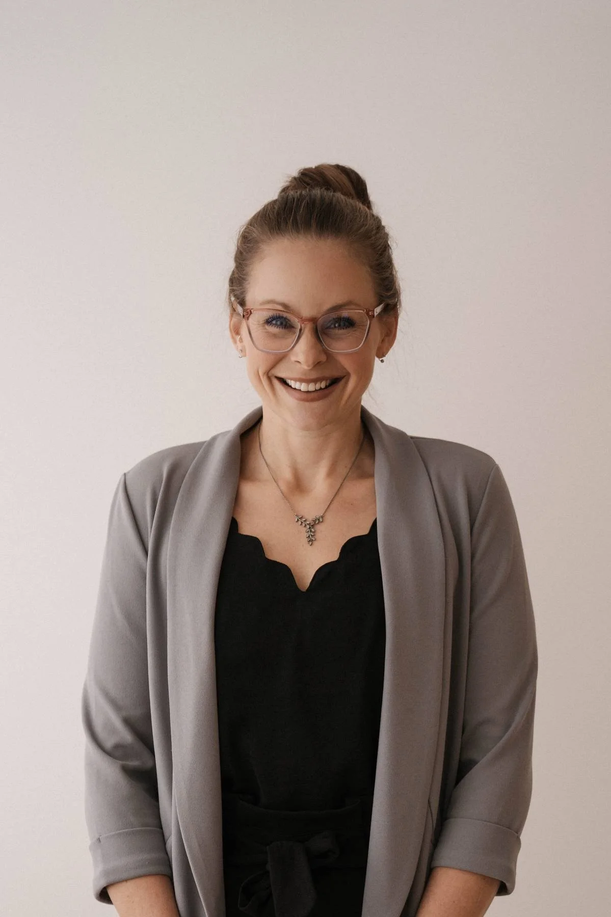 A woman smiling, wearing glasses, a gray blazer, a black top, and a necklace, standing against a plain background.