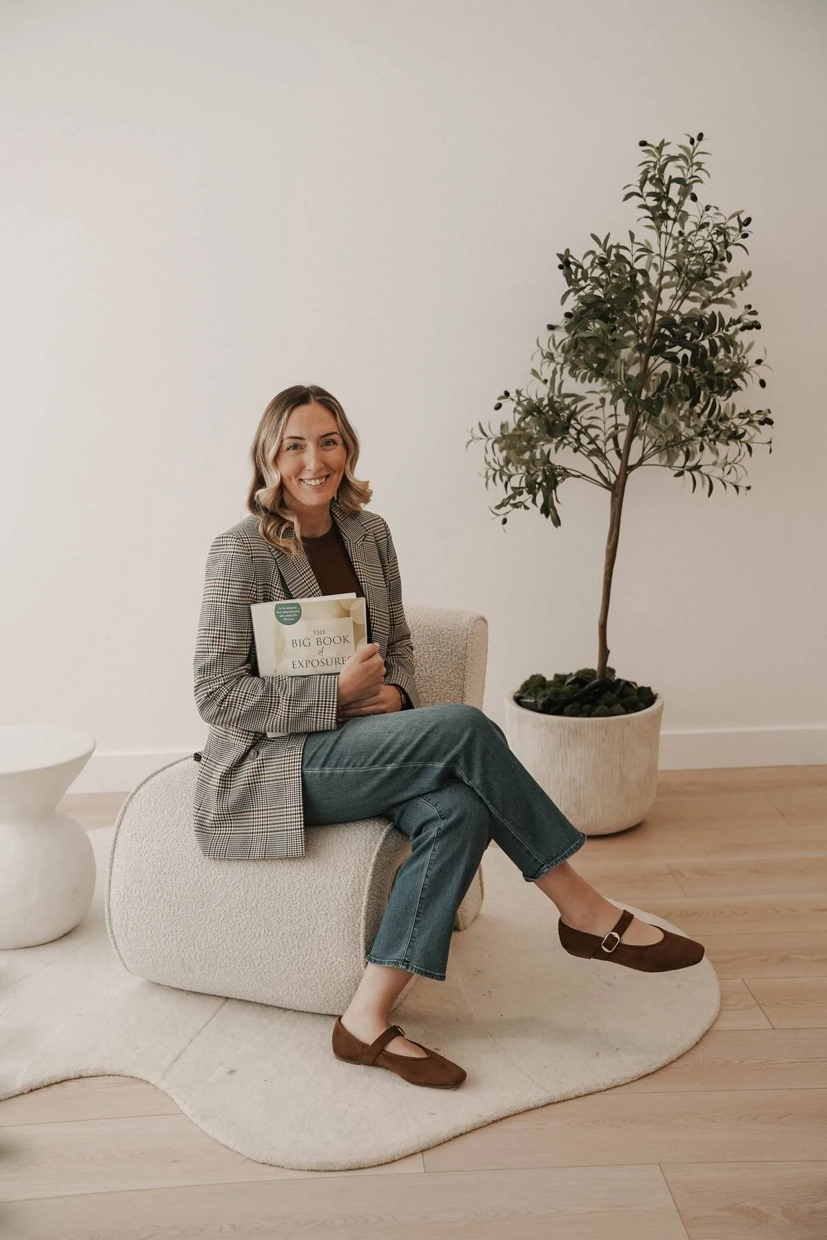 A woman sitting on a beige upholstered chair, holding a book titled 'The Big Book of Exposure,' smiling, with a large potted plant behind her, in a minimalistic room with white walls and wooden flooring.