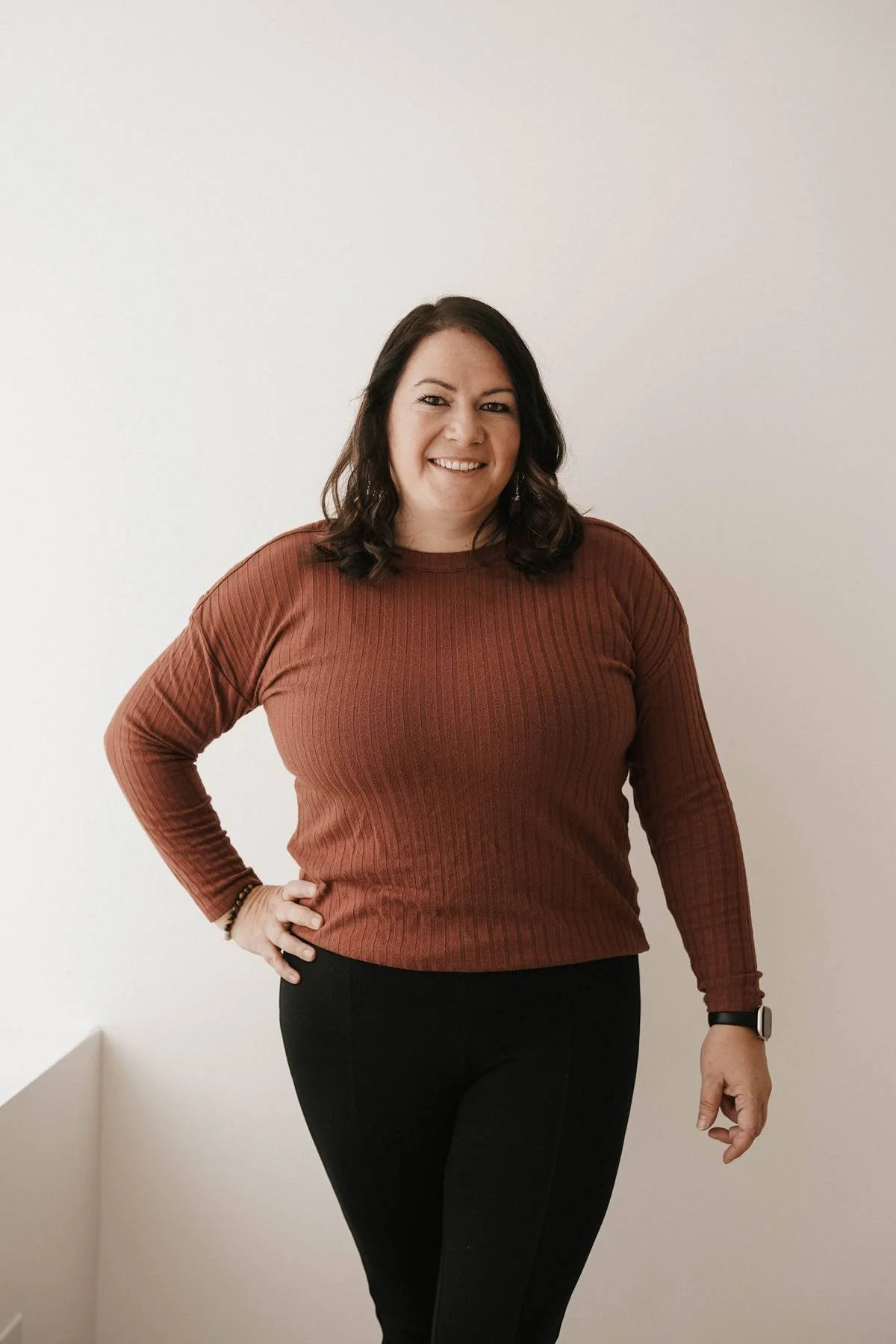 A woman with dark hair, wearing a rust-colored ribbed long-sleeve shirt, black pants, and a smartwatch, standing against a plain white wall with her left hand on her hip and smiling.