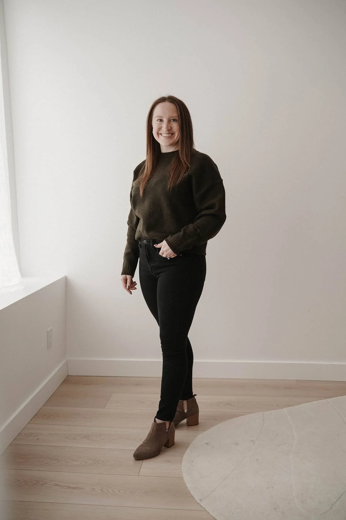 A woman with long red hair smiling and standing in a room with wood floor and white walls, wearing a dark sweater, black jeans, and beige ankle boots.